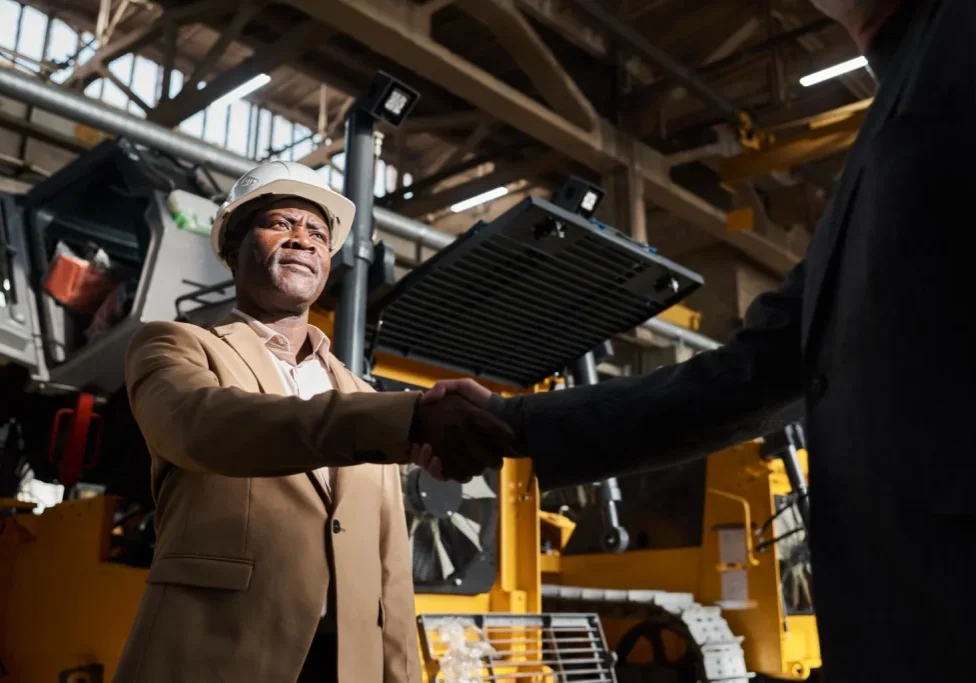 Manufacturing executive and sales partner shaking hands on a factory floor, representing hiring and leadership in industrial manufacturing sales roles.
