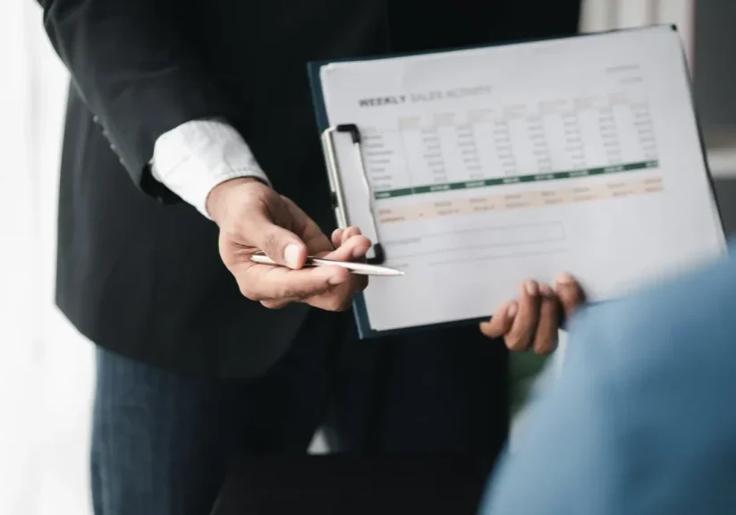 Hiring manager pointing a pen at a weekly sales activity scorecard with daily totals, goal, and variance rows