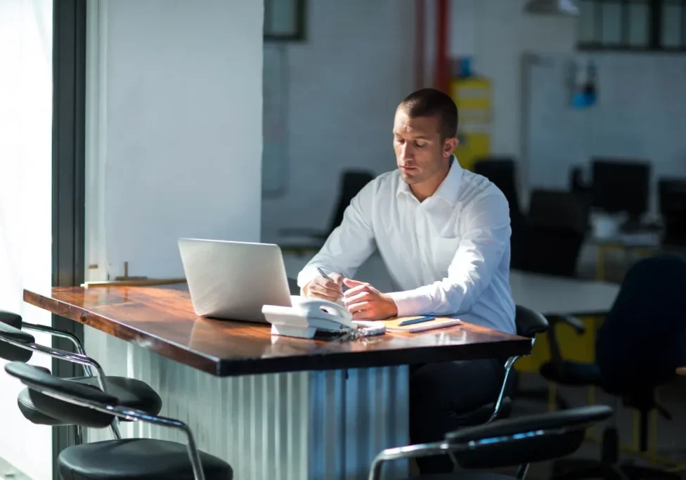 Sales professional in a white shirt taking notes at a wooden desk with a laptop in an industrial office